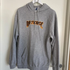 Grå hoodie med No Sense-tryck - Säljer en grå hoodie från No Sense med fet orange och svart logga på bröstet. Tröjan har huva med snörning, stor magficka och långa ärmar. Perfekt för chill dagar och streetwear-stil.