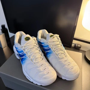 Nike Air Max Plus vita/blå sneakers - Nike Air Max Plus sneakers i vitt och blått med snygga gradientdetaljer och gula loggor. Skorna har en sportig vibe med synliga luftbubblor i sulan, snörning och meshmaterial för extra komfort. Perfekta för dig som vill sticka ut med en fräsch och modern look.