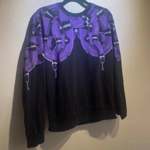 Svart tröja med lila ormtryck Million&Man - Cool svart tröja från Million&Man i storlek S med ett unikt lila ormtryck över axlar och bröst. Tröjan har rund halsringning och långa ärmar. Perfekt statement piece för dig som vill sticka ut med en edgy vibe.
