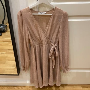 Klänning - Supersöt kort omlottklänning från NA-KD i rosa med diskret blommönster. Klänningen har långa puffärmar, v-ringning och knyt i midjan. Materialet är lätt och har en fin struktur som ger en lyxig känsla. Perfekt för dig som gillar romantisk stil.