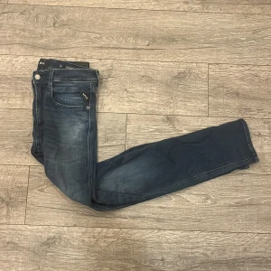 Replay Anbass jeans blå slim fit - Snygga Replay Anbass jeans i mörkblå tvätt med coola slitningar och klassiska fem fickor. Slim fit-modell med normal midja och raka ben. Jeansen har Replay-logga på fickan och metallknapp i midjan. Perfekt för dig som gillar stilrena och moderna jeans. Storleken är 33/32 men sitter mer som W31/32