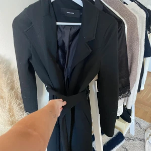 Svart kappa från Vero Moda med bälte - Säljer en svart kappa från Vero Moda med klassisk krage och knytbälte i midjan. Jackan är lång och har en stilren look som passar perfekt till höst och vinter. Materialet är mjukt och känns lyxigt, och kappan har långa ärmar och slät yta.