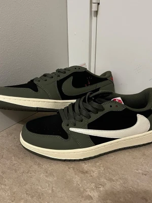 Nike Air Jordan 1 Low Olive/Black - Nike Air Jordan 1 Low med olivgröna och svarta paneler, vit swoosh och röd Air Jordan-logga på hälen. Skorna har snörning, rund tå och platt sula. Materialet är en mix av skinn och syntet, med klassisk Jordan-design och låg siluett.