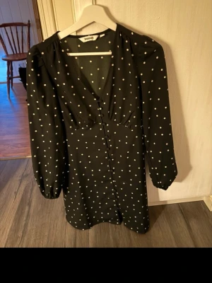 Svart prickig kortklänning Kappahl XS - Supersnygg svart klänning från Kappahl i storlek XS med vita prickar, v-ringad hals och långa ärmar. Klänningen har knappar framtill och en feminin passform som ger en cool vintagekänsla. Perfekt för dig som gillar klassiska mönster och stilrena detaljer.