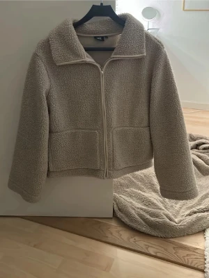 Beige teddyjacka från lager - Mysig beige teddyjacka från lager 157 med krage och dragkedja framtill. Jackan har två stora fickor och är tillverkad i mjukt fleece-material. Perfekt för kyliga dagar och har en avslappnad passform med långa ärmar. 