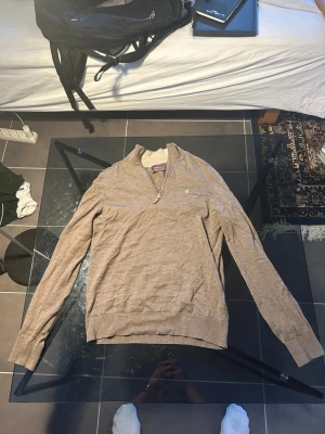 Beige half zip tröja från Morris - Snygg beige långärmad tröja från Morris i 100% merinoull. Tröjan har en stilren half zip med läderdetalj på dragkedjan och ett broderat emblem på bröstet. Perfekt för dig som gillar klassisk och clean stil.