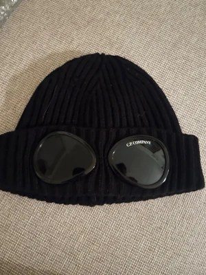 Svart stickad mössa C.P. Company - Säljer en svart ribbstickad mössa från C.P. Company med ikoniska glasögonlinser framtill. Mössan har en cool och unik design som sticker ut och är perfekt för dig som vill ha något extra. Materialet är mjukt och värmande stickat tyg.
