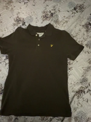  piké från Lyle & Scott - Säljer en olivgrön pikétröja från Lyle & Scott i storlek M. Toppen har klassisk krage, korta ärmar och knappar framtill. Materialet är ekologisk bomull med lite elastan för extra komfort. På bröstet finns Lyle & Scotts ikoniska gula fågelbrodyr. Perfekt för firanden!  