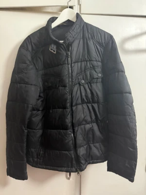 Svart pufferjacka från Barbour XL - Svart pufferjacka från Barbour International i storlek XL. Jackan har quiltad design, ståkrage med spänne, dragkedja framtill och fickor med knappar. Materialet är syntet och jackan är perfekt för kyliga dagar.