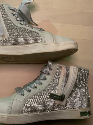 Glittriga vita sneakers från MK - Säljer ett par vita och silverfärgade sneakers från Michael Kors med glittriga stenar på sidorna, silvriga satinband som snörning och dragkedja på sidan. Skorna har rund tå och platt sula, samt MK-detalj framtill. Perfekt för dig som gillar att sticka ut!