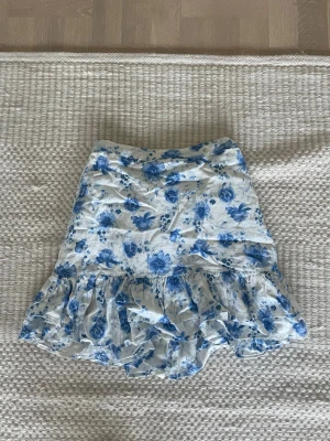 Blommig volangkjol från Lindex - Supersöt kort kjol från Lindex med blått blommönster på vit botten. Kjolen har volangkant nertill och resår i midjan för skön passform. Perfekt för sommarens alla dagar och enkel att matcha med en enkel topp.