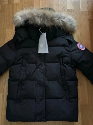 Svart pufferjacka från Canada Goose - Svart pufferjacka från Canada Goose med fluffig päls på huvan och klassisk logga på ärmen. Jackan har dragkedja och knappar framtill, quiltad design och normal passform. Perfekt för kalla vinterdagar och har snygga detaljer som ger en exklusiv vibe.