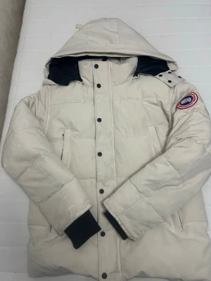 Beige dunjacka från Canada Goose - Säljer en beige dunjacka från Canada Goose med huva och svarta detaljer på insidan. Jackan har ett klassiskt märke på ärmen, svarta knappar och muddar samt två sidofickor. Perfekt för kalla dagar och har en snygg, quiltad look.