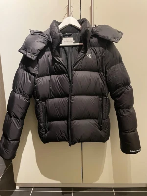 Svart pufferjacka från Calvin Klein Jeans - Säljer en svart dam pufferjacka från Calvin Klein Jeans i storlek L. Jackan är knappt använd och i väldigt fint skick. Den är i storlek L men passar som en M, den är liten i storleken. 