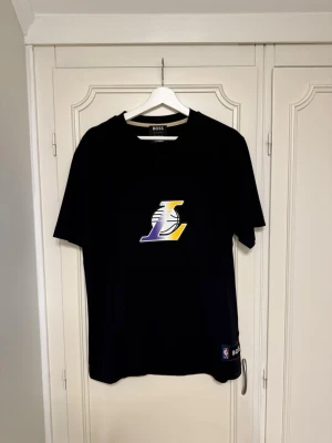 Svart Lakers t-shirt från BOSS - Säljer en svart t-shirt från BOSS i storlek M med Los Angeles Lakers-logga i lila, gult och vitt på bröstet. T-shirten har korta ärmar och rund hals. Perfekt för dig som gillar basket och vill ha en clean och sportig stil. Nästan i nyskick!