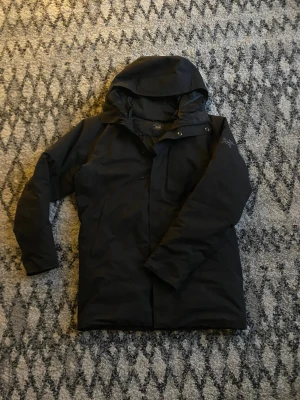 Svart dunjacka från Arc'teryx - Svart dunjacka från Arc'teryx med huva och stilren design. Jackan har broderad logga på ärmen, dragkedja och tryckknappar framtill. Perfekt för kalla dagar och har en normal passform. Materialet är polyester och dun, vilket gör den både varm och lätt.