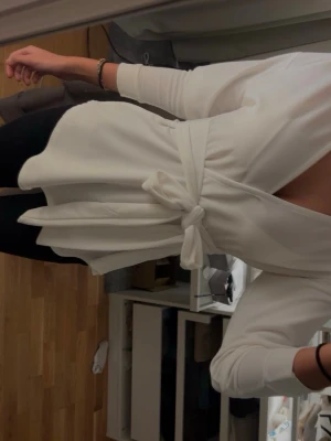 Vit omlottblus med knyt i midjan - Säljer en stilren vit omlottblus med långa ärmar och knyt i midjan. Blusen har v-ringad hals och är tillverkad i ett mjukt, slätt material som ger en snygg siluett. Perfekt att matcha med jeans eller kjol för en fräsch look.