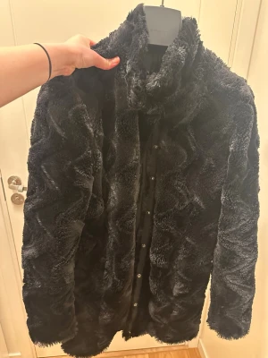 Svart pälsjacka från Vero Moda - Säljer en svart pälsjacka från Vero Moda i storlek L. Jackan har ett vågigt mönster i pälsen, hög krage och knappar framtill. Den är fluffig och har långärmade ärmar, perfekt för att hålla sig varm under kyliga dagar.