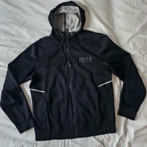 Hugo Boss hoodie  - Svart Hugo Boss hoodie med dragkedja. Jackan har en luva med dragsko och muddar i ärmslut och nederkant. Logotypen är placerad på bröstet. På grund av att  snören gått sönder ( lite, se den sista bilden) säljer billigt.  Nypris minimum 1800 kr