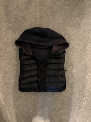 Svart Peak Performance pufferjacka med huva - Svart pufferjacka från Peak Performance med huva och långärmade ärmar i sweatshirt-material. Jackan har quiltad framsida, dragkedja med logotyp och snygg patch på ärmen. Perfekt för dig som gillar sportig och chill stil. Ända defekt är att den är solblek på ena armen och luvan annars superfin.