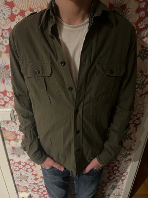 Olivgrön overshirt från Eton - Säljer en olivgrön overshirt från Eton i slim fit. Skjortan har två bröstfickor med knappar, axelklaffar och klassisk krage. Materialet är bomull och den har långa ärmar. Perfekt för dig som gillar stilrena och funktionella plagg.