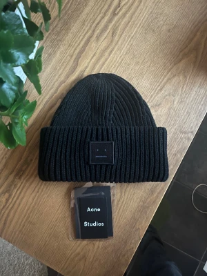 Svart stickad mössa Acne Studios - Säljer en svart stickad mössa från Acne Studios med den ikoniska ansiktsloggan framtill. Mössan har bred uppvikt kant och är tillverkad i mjukt stickat material som håller dig varm. Perfekt accessoar för kalla dagar och snygg till streetwear.