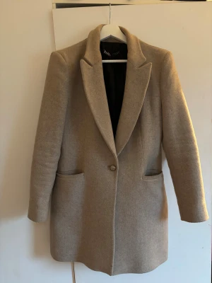 Beige kappa från Zara – klassisk & stilren | Strl M - Stilren och tidlös beige/kamel kappa från Zara i figurnära, klassisk modell. Rak passform med fodrade axlar, slagkrage, fickor och en knapp framtill – passar både vardag och mer uppklätt. • Märke: Zara • Storlek: M • Färg: Beige/camel • Skick: Väldigt bra skick • Notering: Mindre slitage i fodret invändigt (se bild), ej synligt vid användning Lätt att styla och en riktig garderobsklassiker ✨
