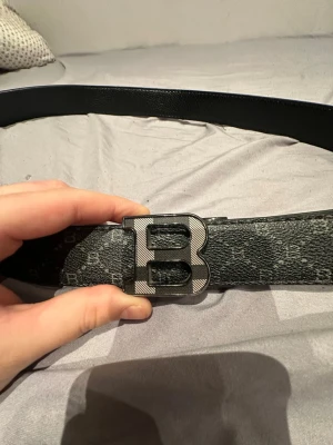 Svart skärp med B-spänne - Snyggt svart skärp från Balmain med ett stort B-format metallspänne. Skärpet har ett mönster med små B och prickar över hela ytan och är gjort i präglat skinn. Perfekt för att lyfta din outfit med en exklusiv touch.