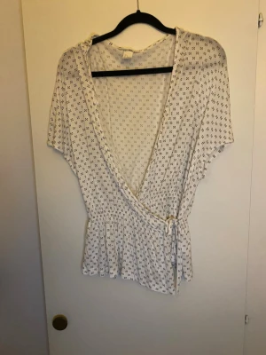 Vit omlottblus med små blommor - Säljer en vit omlottblus med små svarta blommor, v-ringad och med knyt i midjan. Blusen har korta ärmar och ett mjukt, lätt material som känns skönt mot huden. Perfekt för vår och sommar, med en feminin volangkant nedtill. En knapp sitter lös
