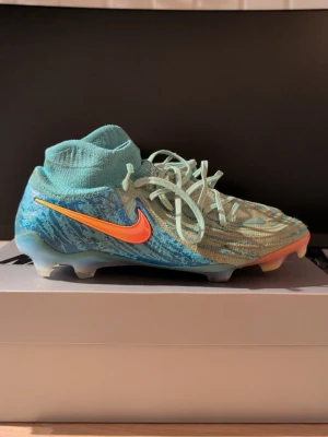 Nike Phantom fotbollsskor blå/turkos - Nike Phantom fotbollsskor i blå och turkos med orange swoosh. Skorna har snörning, stickad ovandel och platt sula med dobbar för gräsplan. Mönstrad design med inslag av ljusblått och grönt, samt detaljer i gult och rosa på sulan. Perfekt för dig som vill sticka ut på planen.