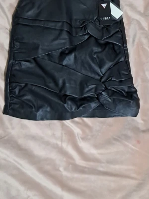 Svart kort kjol i skinn från Guess - Säljer en svart kort kjol från Guess i snyggt skinnmaterial. Kjolen har coola draperade detaljer framtill och en synlig dragkedja bak med Guess-logga på zip-pullern. Passformen är normal och kjolen har en stilren look som är lätt att matcha. Xs/S
