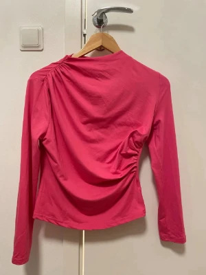Rosa långärmad topp med rynk - Säljer en snygg rosa långärmad topp med coola rynkdetaljer på axeln och sidan. Toppen har en rund hals och är gjord i mjukt bomullsmaterial. Perfekt för dig som gillar färg och vill ha något som sticker ut lite extra.💕