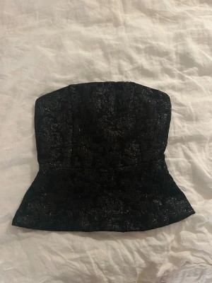 Svart glittrig topp - Säljer en svart tub topp med glittrig yta och struktur i tyget. Toppen är axelbandslös och har en figurnära passform som markerar midjan. Den är skit snygg speciellt när man ska klä upp sig. Använd en gång