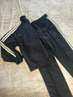 Svarta träningskläder från Nova Men - Säljer ett svart träningsset från Nova Men med vita ränder längs ärmar och ben. Jackan har dragkedja och ribbade muddar, byxorna har elastisk midja och raka ben. Materialet är mjukt och syntetiskt, perfekt för sport eller chill.