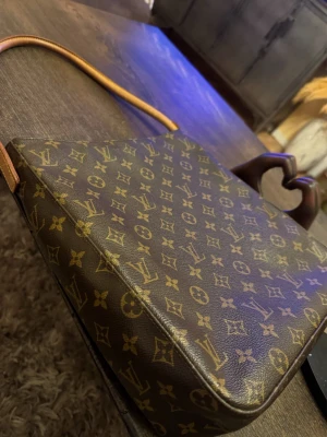 Louis Vuitton brun axelväska monogram - Säljer en klassisk brun axelväska från Louis Vuitton med ikoniskt monogrammönster i guld. Väskan har en lång rem i skinn och dragkedja upptill. Insidan är ljus och har en ficka för småsaker. Perfekt storlek för det viktigaste och riktigt snygg design.