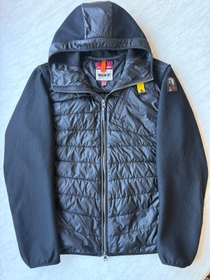 Parajumpers cardigan - svart cardigan jacka från Parajumpers med huva och coolt gult spänne vid halsen. Jackan har quiltad framsida, mesh-aktiga ärmar och bröstficka med dragkedja. Märkets patch på ena ärmen ger extra stilpoäng. Perfekt för dig som gillar street och funktion. (Storlek L) för fler bilder eller frågor hör av dig.