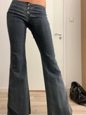 Mörkblå bootcut jeans från Ex-It - Snygga mörkblå bootcut jeans från Ex-It med låg midja och tre dekorativa knappar framtill. Jeansen har en bred linning och är tillverkade i ett stretchigt jeansmaterial som sitter bekvämt. Perfekt för dig som gillar retrovibbar och vill ha ett statement-plagg.