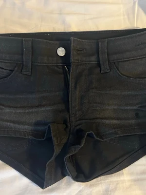 Svarta shorts från Cheap Monday - Säljer ett par svarta shorts från Cheap Monday med klassisk femficksdesign och grönt logopatch baktill. De har låg midja, dragkedja och knapp framtill samt bälteshällor. Materialet är bomull med lite stretch för skön passform.