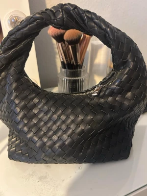 Svart flätad handväska i skinn - Säljer en svart handväska med snygg flätad design och rundat handtag. Väskan är tillverkad i skinn och har en praktisk dragkedja upptill. Perfekt storlek för att få plats med det viktigaste och ger en cool vibe till din outfit.