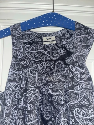 Svart paisleymönstrad klänning Acne studios  - Säljer en svart ärmlös klänning från Acne med vitt paisleymönster och blommor. Modellen är rak och knälång med rund asymmetrisk  halsringning . Materialet känns lätt och luftigt, perfekt för dig som gillar unika prints och stilren design.