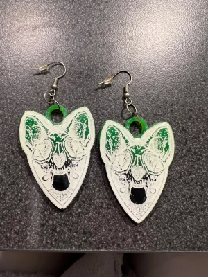 Grön svarta skelett örhängen  - Unik örhängen i grönt och svart med coolt tryck av ett djurkranium på framsidan. Örhängena har en spetsig form nertill och med vita detaljer. Designen är riktigt edgy och passar dig som gillar att sticka ut med din stil.