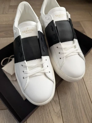 Valentino Garavani Open ev byte - Säljer ett par Valentino Garavani Open sneakers i vitt skinn med bred svart rand över mitten. Klassisk rund tå, platt sula och vita snören. 👌 kan även bytas mot förslag!