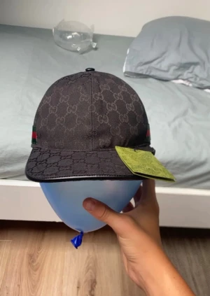 Svart Gucci keps med GG-mönster - Säljer en svart Gucci keps med klassiskt GG-mönster över hela kepsen. Kepsen har en snygg grön och röd rand på sidan och är tillverkad i bomullsmaterial. Justerbar baktill med metallspänne. Perfekt för dig som vill ha en lyxig streetstyle-accessoar.