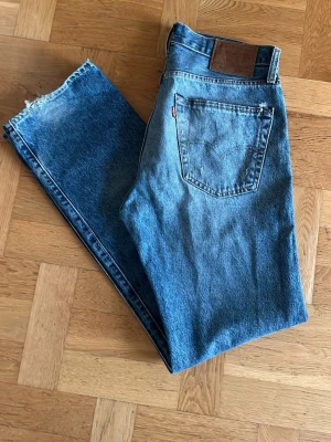 Levi's 501 blå jeans - Klassiska Levi's 501 jeans i blå denim med raka ben och femficksdesign. Jeansen har en brun läderpatch bak i midjan och den ikoniska röda Levi's-taggen på bakfickan. Perfekt för dig som gillar tidlös stil och äkta denim.