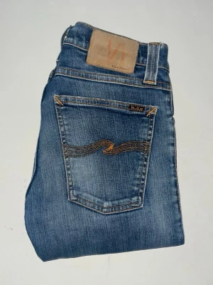 Nudie Jeans - Säljer ett par klassiska blå skinny jeans från Nudie Jeans, i modellen tajht long john. Jeansen har snygga slitningar och kontrastsömmar. Midja 33cm, längd 106cm. JAG SKICKAR EJ FLER BILDER NÄR JAG BÄR JEANSEN! för bättre inblick i passformen rekommenderar jag att googla på modellnamnet. För storleksguide kolla måtten noggrant då jeansen alltid kan vara uppsydda heller krympta och jag ej tar emot returer :) s5,2