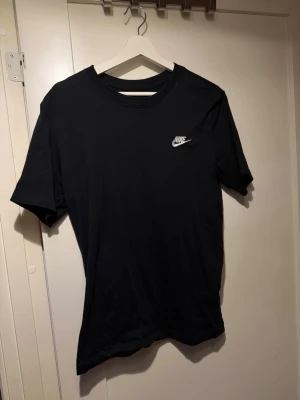 Svart Nike t-shirt i bomull - Klassisk svart t-shirt från Nike med vit logga på bröstet. Skön passform och tillverkad i mjuk bomull. Perfekt till jeans eller shorts för en clean och sportig look.