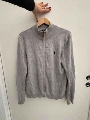 Grå kofta från Polo Ralph Lauren - Snygg grå kofta från Polo Ralph Lauren i bomull med dragkedja och ribbad krage. Klassisk logga broderad på bröstet och diskreta muddar vid ärmslut och nederkant. Perfekt som julklapp. Köpt för 1200kr pris kan diskuteras.