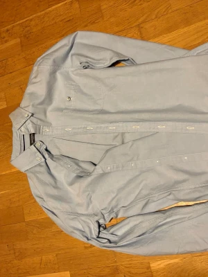 Ljusblå skjorta från Dressmann - Snygg ljusblå skjorta från Dressmann med klassisk krage och knappstängning hela vägen. Skjortan har en bröstficka med knapp och är långärmad. Tillverkad i mjuk bomull som känns skön mot huden. Perfekt för dig som gillar stilrena och enkla plagg. Helt by med prislappen kvar!