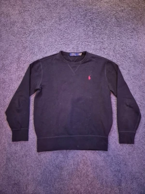 Svart Polo Ralph Lauren sweatshirt - Svart sweatshirt från Polo Ralph Lauren med klassisk rund hals och klassisk långärmad design. Tröjan har en röd broderad logga på bröstet och är tillverkad i mjuk bomull. Passformen är normal och den har ribbade muddar vid ärmslut och nederkant. Givetvis så är den äkta och de är bara att höra av er vid frågor😁!