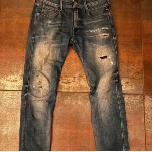 Replay Anbass med slitningar - Säljer ett par Replay Anbass jeans i blå tvätt med slitna detaljer och snygga hål på benen. Modellen är skinny med normal passform och klassiska fem fickor. Jeansen har dragkedja och är gjorda i stretchig bomull för skön känsla.